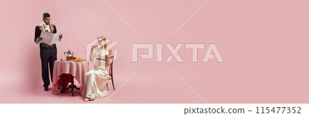 Man in velvet jacket and cravat reads newspaper while woman in satin gown and gloves drinks tea at table against pink studio background Man in velvet jacket and cravat reads newspaper while woman in satin gown and gloves drinks tea at table against pink studio background 115477352