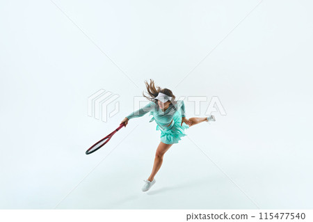 Young woman, tennis player in mint-green outfit, mid-serve, with ball and racquet in action against white studio background. Young woman, tennis player in mint-green outfit, mid-serve, with ball and racquet in action against white studio background. 115477540