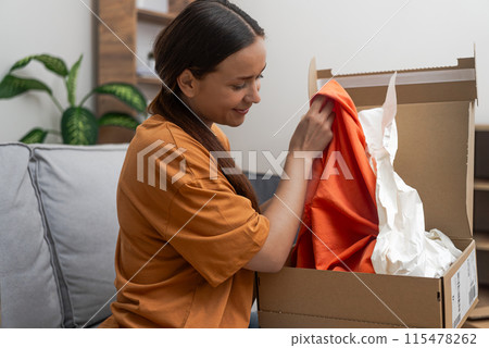 woman on her sofa examines her express delivery, expressing delight after a satisfying e-commerce fashion spree. 115478262