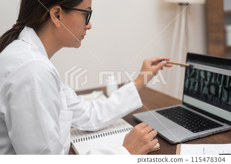 X-ray Diagnosis: Focused and analytical, a young doctor in glasses analyzes X-ray images displayed on a laptop. X-ray Diagnosis: Focused and analytical, a young doctor in glasses analyzes X-ray images displayed on a laptop. 115478304