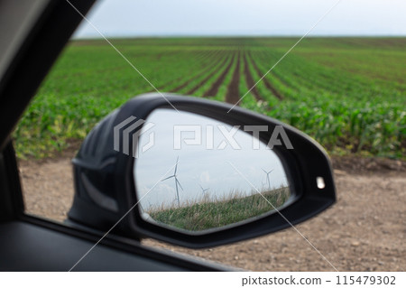 Windmill to produce electricity in cars mirror, behind green plantations grow in the field 115479302