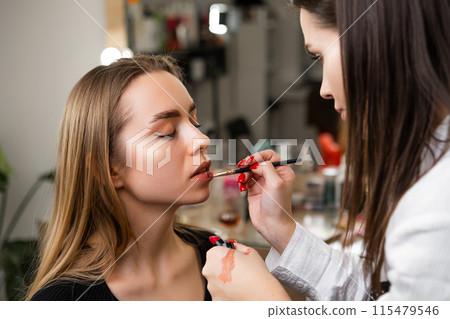 Makeup artist applies lipstick with gentle strokes using special brush. Use of brush provides artist with control over woman lips edges Makeup artist applies lipstick with gentle strokes using special brush. Use of brush provides artist with control over woman lips edges 115479546