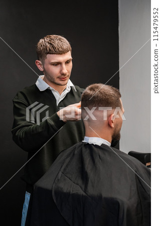 Young barber carefully cuts the mans hair with a trimmer in the bustling barbershop Young barber carefully cuts the mans hair with a trimmer in the bustling barbershop 115479552