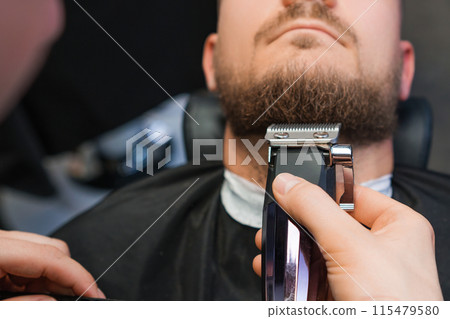 Master skillfully trims the clients beard using an automatic trimmer in the barbershop 115479580