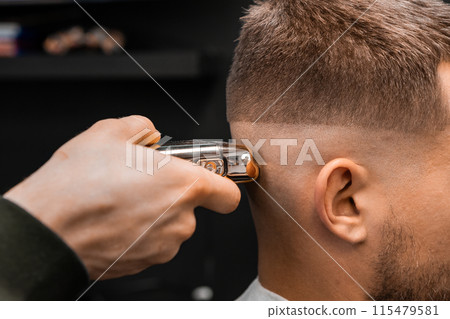 Barber uses shaver machine to cut young man hair in barbershop closeup. Master does modern haircut to male client in professional salon Barber uses shaver machine to cut young man hair in barbershop closeup. Master does modern haircut to male client in professional salon 115479581