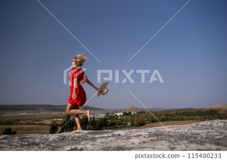 A woman in a red dress is running in a field with a straw hat on 115480233
