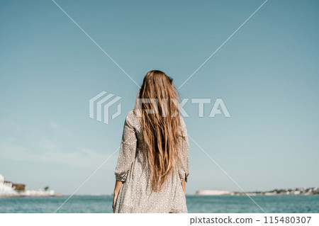 woman with long hair stands in front of a blue sky. She is wearing a white and black striped shirt. 115480307