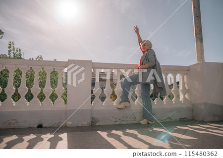 A woman in a gray jacket and blue jeans is leaning against a white railing. She is holding a book in her hand. The scene has a calm and relaxed mood, with the woman enjoying a quiet moment outdoors. 115480362