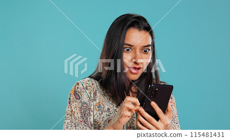 Woman happily scrolling on phone touchscreen to check social media feed, amazed by photo. Indian person browsing internet websites, astonished by content, studio background, camera B Woman happily scrolling on phone touchscreen to check social media feed, amazed by photo. Indian person browsing internet websites, astonished by content, studio background, camera B 115481431