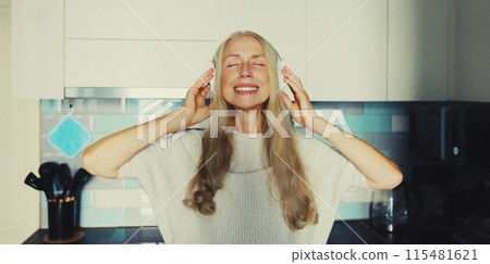 Happy smiling mature woman in headphones enjoying music with closed eyes in the kitchen at home Happy smiling mature woman in headphones enjoying music with closed eyes in the kitchen at home 115481621