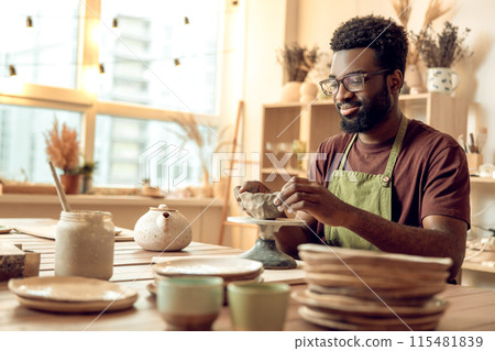 Curly-haired man sitting at the table, working with clay and looking involved Curly-haired man sitting at the table, working with clay and looking involved 115481839