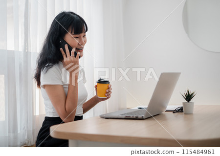 A woman is talking on her cell phone while holding a coffee cup. She is sitting at a desk with a laptop and a potted plant. Concept of productivity and multitasking A woman is talking on her cell phone while holding a coffee cup. She is sitting at a desk with a laptop and a potted plant. Concept of productivity and multitasking 115484961