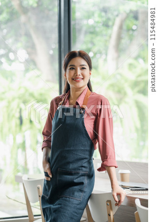 A woman in a blue apron stands in front of a table with a cup of coffee. She is smiling and she is happy A woman in a blue apron stands in front of a table with a cup of coffee. She is smiling and she is happy 115484981