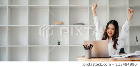 Happy young woman raising arms in celebration while working on a laptop in a modern office with white shelves. Happy young woman raising arms in celebration while working on a laptop in a modern office with white shelves. 115484990