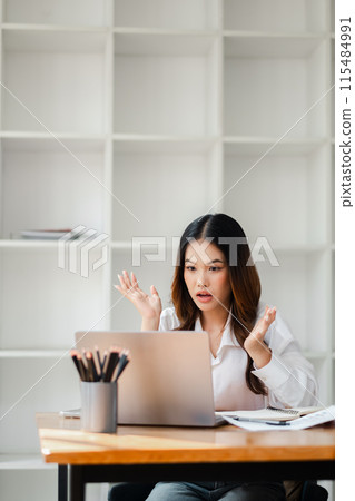 A woman is sitting at a desk with a laptop in front of her. She is looking at the screen with a shocked expression on her face 115484991