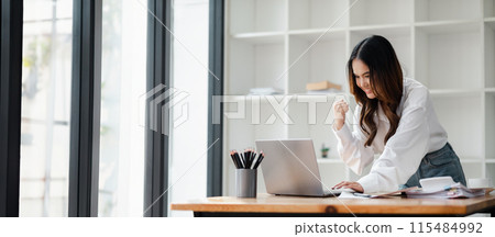 A young professional woman in a modern office, celebrating success while working on her laptop. Bright and contemporary workspace with large windows and shelves in the background. A young professional woman in a modern office, celebrating success while working on her laptop. Bright and contemporary workspace with large windows and shelves in the background. 115484992