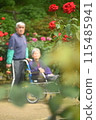 An elderly man in a wheelchair walking in the park with a caregiver assisting him 115485941