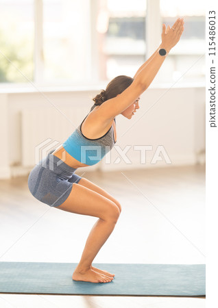 Sportive girl practicing chair pose of yoga in light room Sportive girl practicing chair pose of yoga in light room 115486013