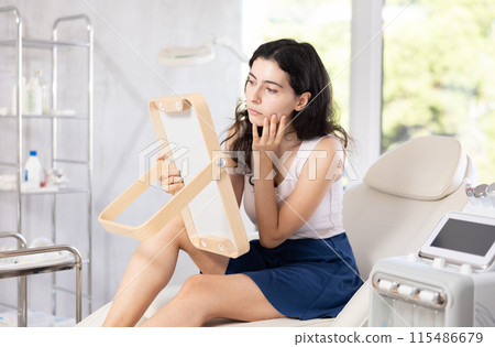 Young brunette woman admires her new face in mirror after plastic surgery 115486679