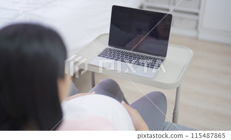 A portrait of happy Asian pregnant woman using, working online with computer laptop notebook tablet device with screen in bedroom at home. Having a baby. Family people lifestyle. Mom love. 115487865