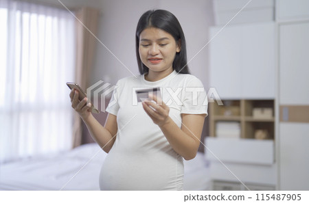 A portrait of happy Asian pregnant woman using a mobile phone device with credit card, shopping online in bedroom at home. Having a baby. Family people lifestyle. Mom love. A portrait of happy Asian pregnant woman using a mobile phone device with credit card, shopping online in bedroom at home. Having a baby. Family people lifestyle. Mom love. 115487905
