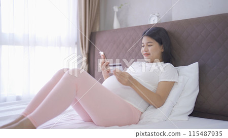 A portrait of happy Asian pregnant woman using a mobile phone device with credit card, shopping online in bedroom at home. Having a baby. Family people lifestyle. Mom love. A portrait of happy Asian pregnant woman using a mobile phone device with credit card, shopping online in bedroom at home. Having a baby. Family people lifestyle. Mom love. 115487935