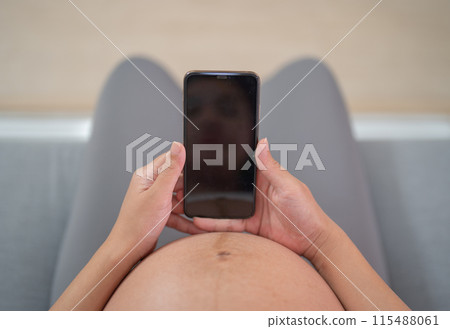 A portrait of happy Asian pregnant woman using online mobile phone technology device with screen in bedroom at home. Having a baby. Family people lifestyle. Mom love. A portrait of happy Asian pregnant woman using online mobile phone technology device with screen in bedroom at home. Having a baby. Family people lifestyle. Mom love. 115488061