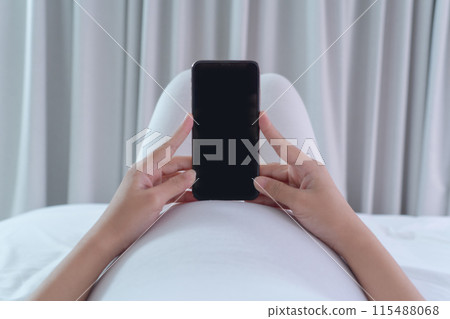 A portrait of happy Asian pregnant woman using online mobile phone technology device with screen in bedroom at home. Having a baby. Family people lifestyle. Mom love. A portrait of happy Asian pregnant woman using online mobile phone technology device with screen in bedroom at home. Having a baby. Family people lifestyle. Mom love. 115488068