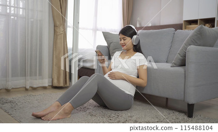 A portrait of happy Asian pregnant woman using online mobile phone technology device with screen in bedroom at home. Having a baby. Family people lifestyle. Mom love. A portrait of happy Asian pregnant woman using online mobile phone technology device with screen in bedroom at home. Having a baby. Family people lifestyle. Mom love. 115488104