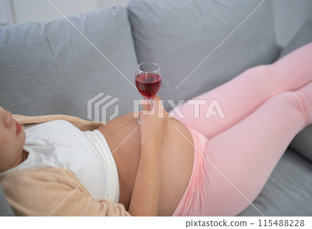 A portrait of happy Asian pregnant woman drinking wine alcohol beverage glass in bedroom at home. Having a baby. Family people lifestyle. Mom. 115488228