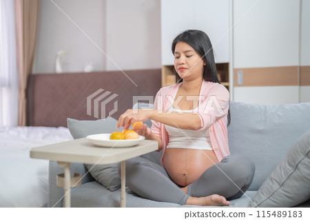 A Portrait of happy Asian pregnant woman eating orange fruit in bedroom at home. Having a baby. Family people lifestyle. Mom love. 115489183