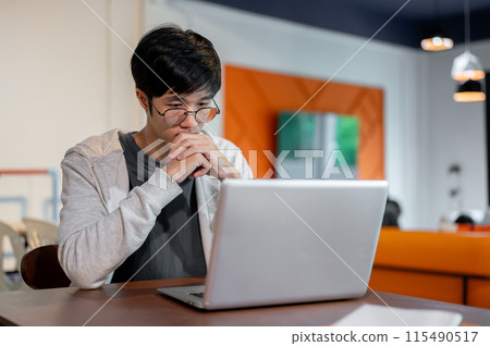A thoughtful young Asian man is working on his laptop, looking at the screen with a serious face. 115490517