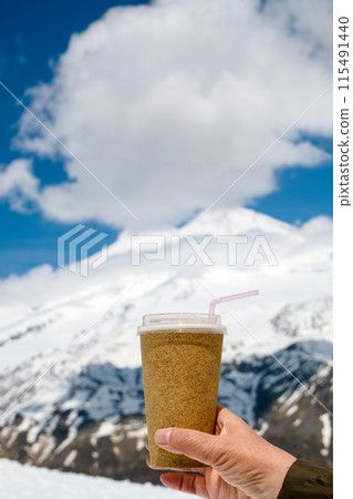 A disposable cup with a drink and a straw in hand against the backdrop of snow-capped mountains and a blue sky with clouds. Vacation in the mountains, hike, travel A disposable cup with a drink and a straw in hand against the backdrop of snow-capped mountains and a blue sky with clouds. Vacation in the mountains, hike, travel 115491440
