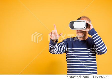 Portrait Asian smiling old man pointing at something in a virtual reality glasses headset studio shot isolated yellow background, Excited happy senior man pensioner using VR play simulation game 115492258