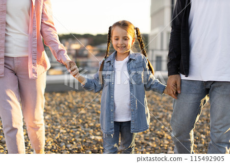 Happy child holding parents' hands during outdoor walk at sunset Happy child holding parents' hands during outdoor walk at sunset 115492905