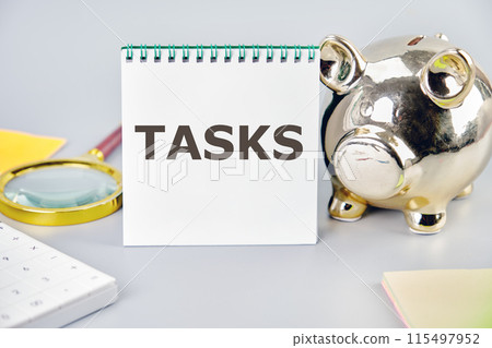 Goals achievement and business success. TASK word on a blank sheet in a notebook in a composition with a piggy bank, magnifying glass, calculator, stickers on a gray background 115497952