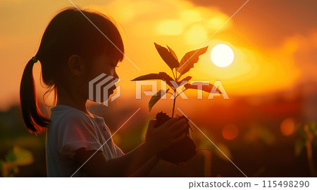 Silhouette of a young Asian girl tenderly holds a young plant against the backdrop of a breathtaking sunset 115498290