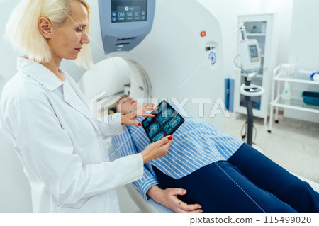 Female patient is lying at CT bed and listening to doctor. Caucasian female doctor looking at tablet with film of MRI tomography scan of the brain. 115499020