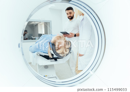Radiographer operating computed tomography scanner being used to scan lying patient in radiology department of hospital. Doctor holding a tablet and looking at the patient. Radiographer operating computed tomography scanner being used to scan lying patient in radiology department of hospital. Doctor holding a tablet and looking at the patient. 115499033