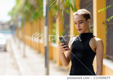 Queer non-binary man with mustache wearing lipstick and jumpsuit using mobile phone outdoors 115499077