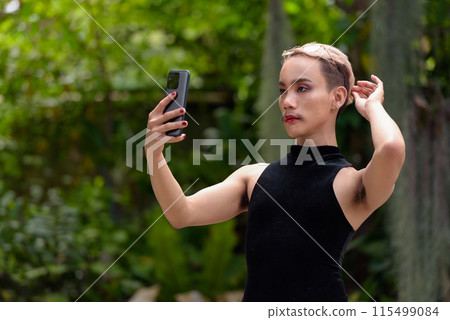 Queer LGBT community supporter man with mustache wearing jumpsuit using phone outdoors in park Queer LGBT community supporter man with mustache wearing jumpsuit using phone outdoors in park 115499084