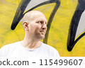 Bald man in white t-shirt looking sideways, colorful graffiti background, urban setting 115499607