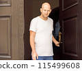 Bald man in white t-shirt holding door handle, standing at entrance, looking forward, serious expression 115499608