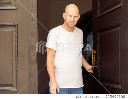 Bald man in white t-shirt holding door handle, standing at entrance, looking forward, serious expression Bald man in white t-shirt holding door handle, standing at entrance, looking forward, serious expression 115499608