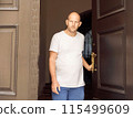 Bald man in white t-shirt holding door handle, looking seriously, outdoor urban setting 115499609