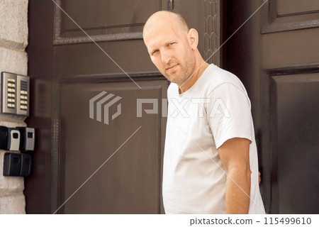 Bald man in white t-shirt holding door handle, standing at entrance, looking forward Bald man in white t-shirt holding door handle, standing at entrance, looking forward 115499610