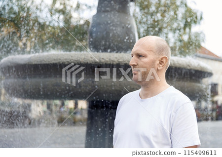 Bald man in white t-shirt standing near fountain, looking thoughtful, outdoor setting 115499611