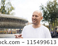 Bald man in white t-shirt smiling, pointing near fountain, outdoor park setting 115499613