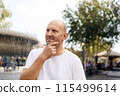 Bald man in white t-shirt smiling, pointing near fountain, outdoor park setting 115499614