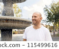 Bald man in white t-shirt smiling, pointing near fountain, outdoor park setting 115499616